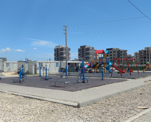 adiyaman11 Konteynerlerin arasında yer alan açık alan; çocuk oyun grupları ve açık hava spor aletleri ile birlikte çevrede yükselen yeni konut inşaatlarının göründüğü geniş bir meydan.