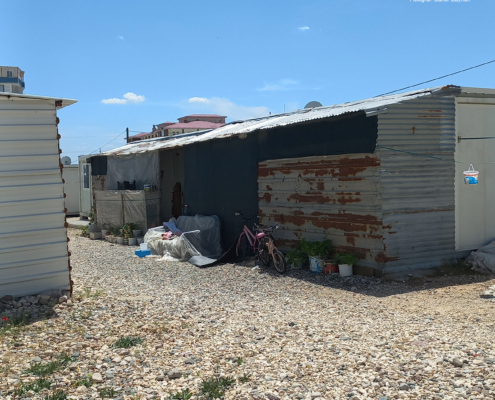 adiyaman13 Dış yüzeyi eski ve yamalı sac levhalarla kaplı bir konteyner; önünde bisiklet, kapitone örtüler ve çeşitli saksılarla birlikte taşlık bir zemin üzerinde kurulu yaşam alanı.