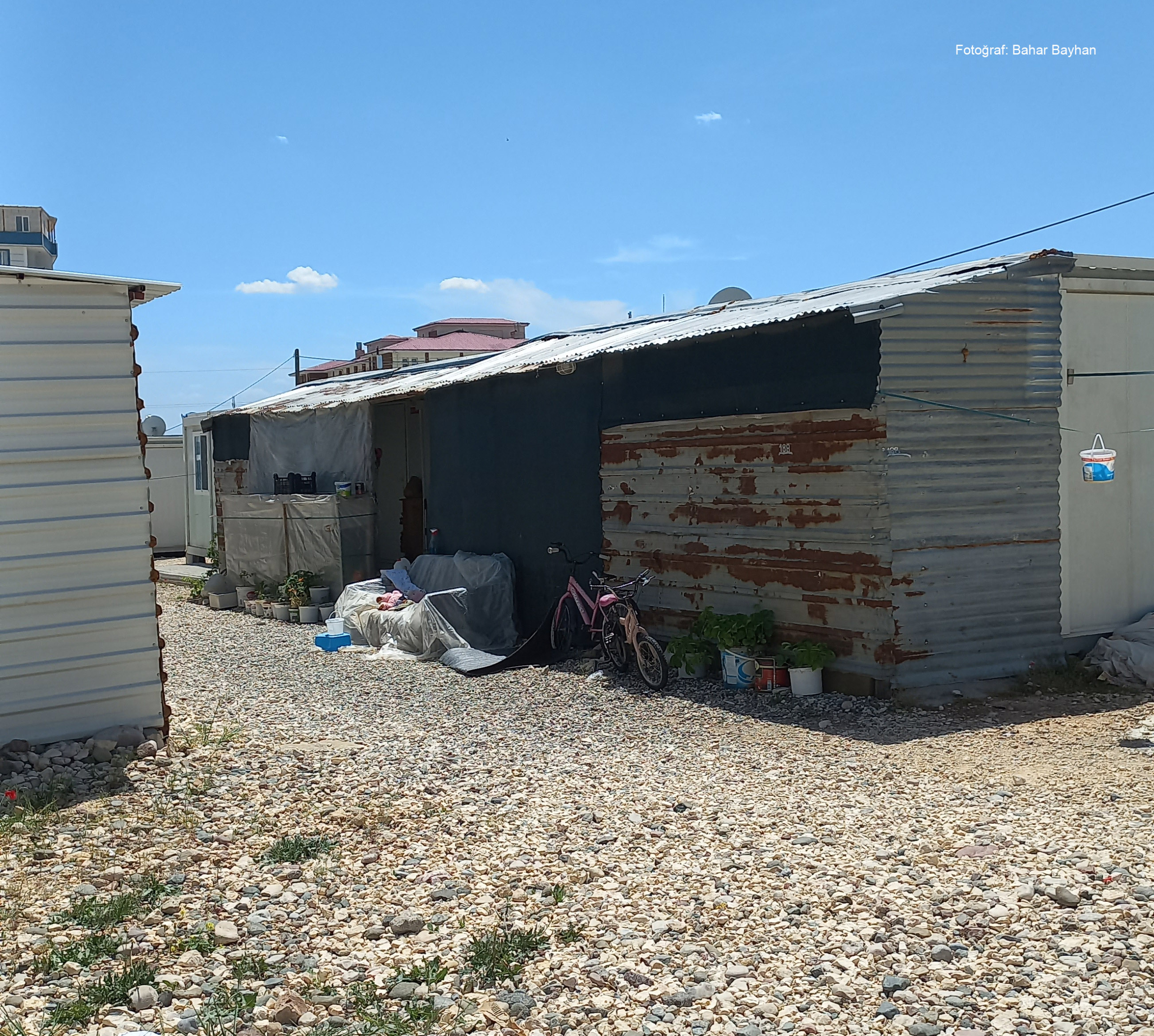 adiyaman13 Dış yüzeyi eski ve yamalı sac levhalarla kaplı bir konteyner; önünde bisiklet, kapitone örtüler ve çeşitli saksılarla birlikte taşlık bir zemin üzerinde kurulu yaşam alanı.