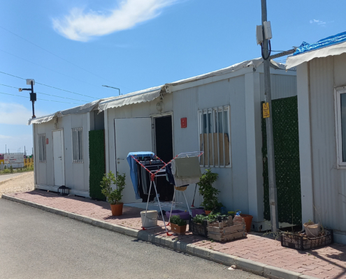 adiyaman14 Beyaz renkli yan yana dizilmiş konteynerler, önünde çamaşırlık, saksı bitkileri ve günlük yaşam düzenlemeleri bulunan bir sokak görünümü.