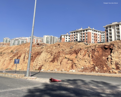 adiyaman3 Yol kenarında yüksek bir şevle ayrılmış bir alanda yükselen yeni apartman blokları; ön planda keskin yüzeyli kırmızımsı kaya kütlesi ve bir sokak lambası yer alıyor.
