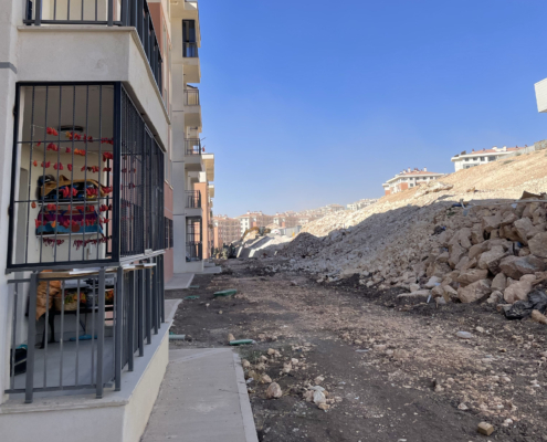 adiyaman4 Yeni yapılmış apartman bloklarının yanında, kazı çalışmaları nedeniyle oluşmuş geniş bir moloz yığını ve düzensiz toprak zemin; ön planda demir korkuluklu bir balkon ve içeride kurutulmuş biberlerin asılı olduğu görülüyor.