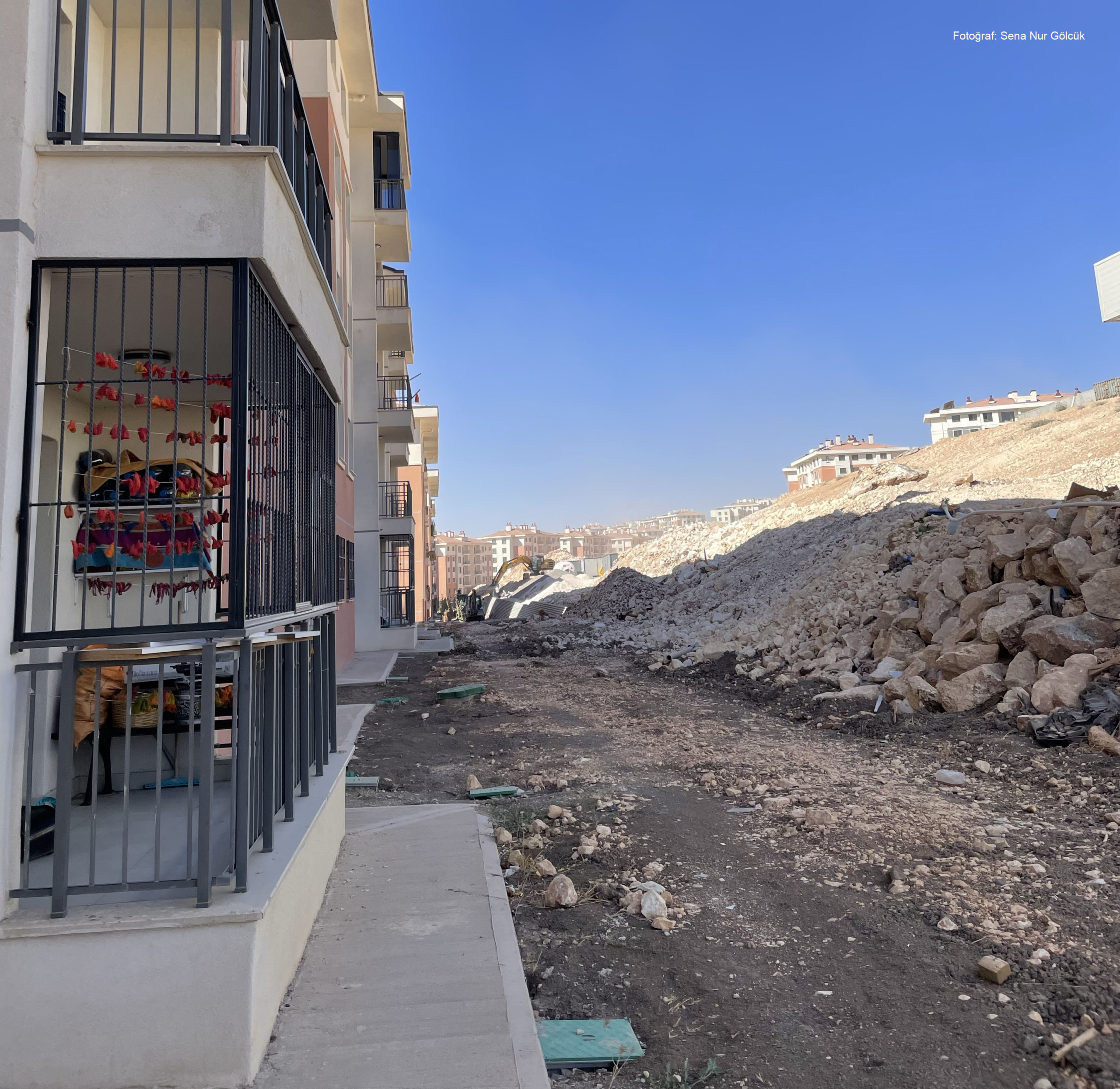 adiyaman4 Yeni yapılmış apartman bloklarının yanında, kazı çalışmaları nedeniyle oluşmuş geniş bir moloz yığını ve düzensiz toprak zemin; ön planda demir korkuluklu bir balkon ve içeride kurutulmuş biberlerin asılı olduğu görülüyor.