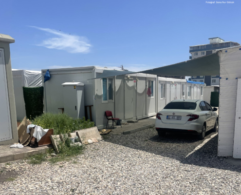 adiyaman7 Konteynerların arasındaki sokakta bir otomobil park etmiş. İki konteyner arasına gölgelik çekilmiş ve araç gölgede duruyor.