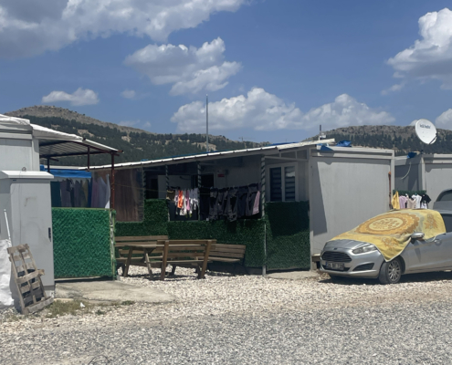 adiyaman8 Konteynerlerin arasına yerleştirilmiş banklar, çamaşır telleri ve suni çitlerle çevrelenmiş küçük bir açık oturma alanı; yanında branda ile örtülmüş bir otomobil.