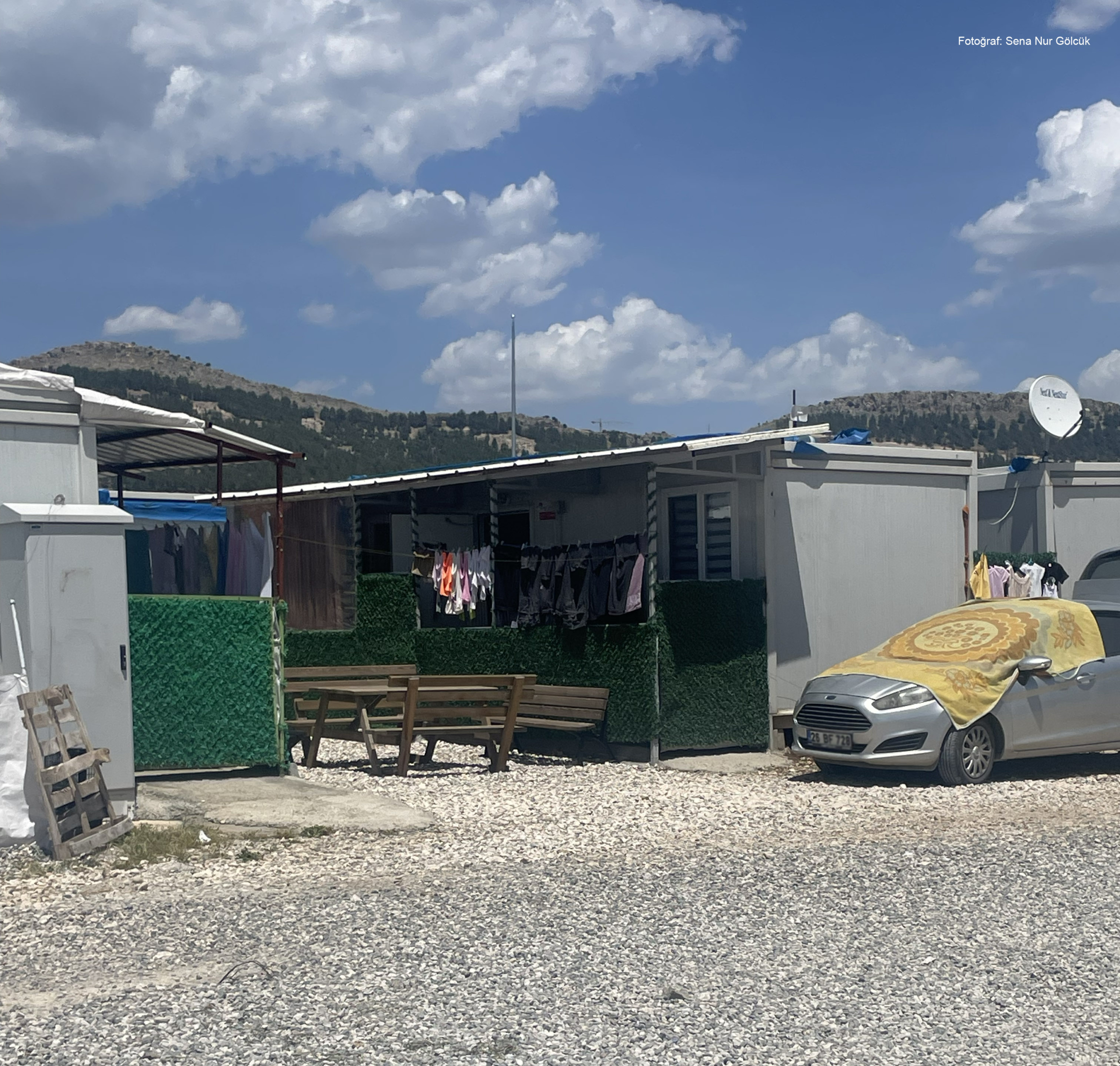 adiyaman8 Konteynerlerin arasına yerleştirilmiş banklar, çamaşır telleri ve suni çitlerle çevrelenmiş küçük bir açık oturma alanı; yanında branda ile örtülmüş bir otomobil.